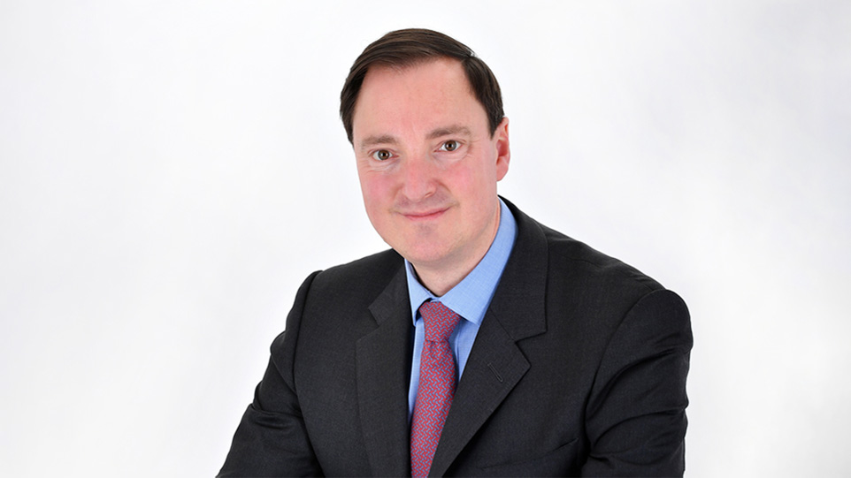 A man with dark hair, wearing a blue shirt and suit, smiling at the camera.